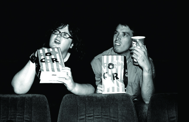 A black and white photo of Nicko and Joe sitting in a cinema, eating popcorn and looking bewildered.