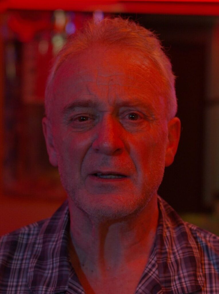 A close up of an older man wearing a checked shirt, bathed in red light.