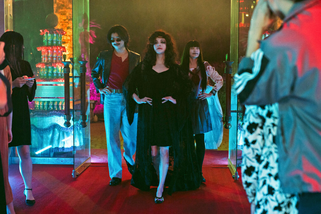 A man, a woman and a girl stride confidently into a room together, their outfits somewhat costume-y. The woman at the centre wears a long gothic dress, and the girl to her left has skull make-up and is wearing a school uniform.