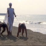 A woman in white holds two naked people on leads on the beach