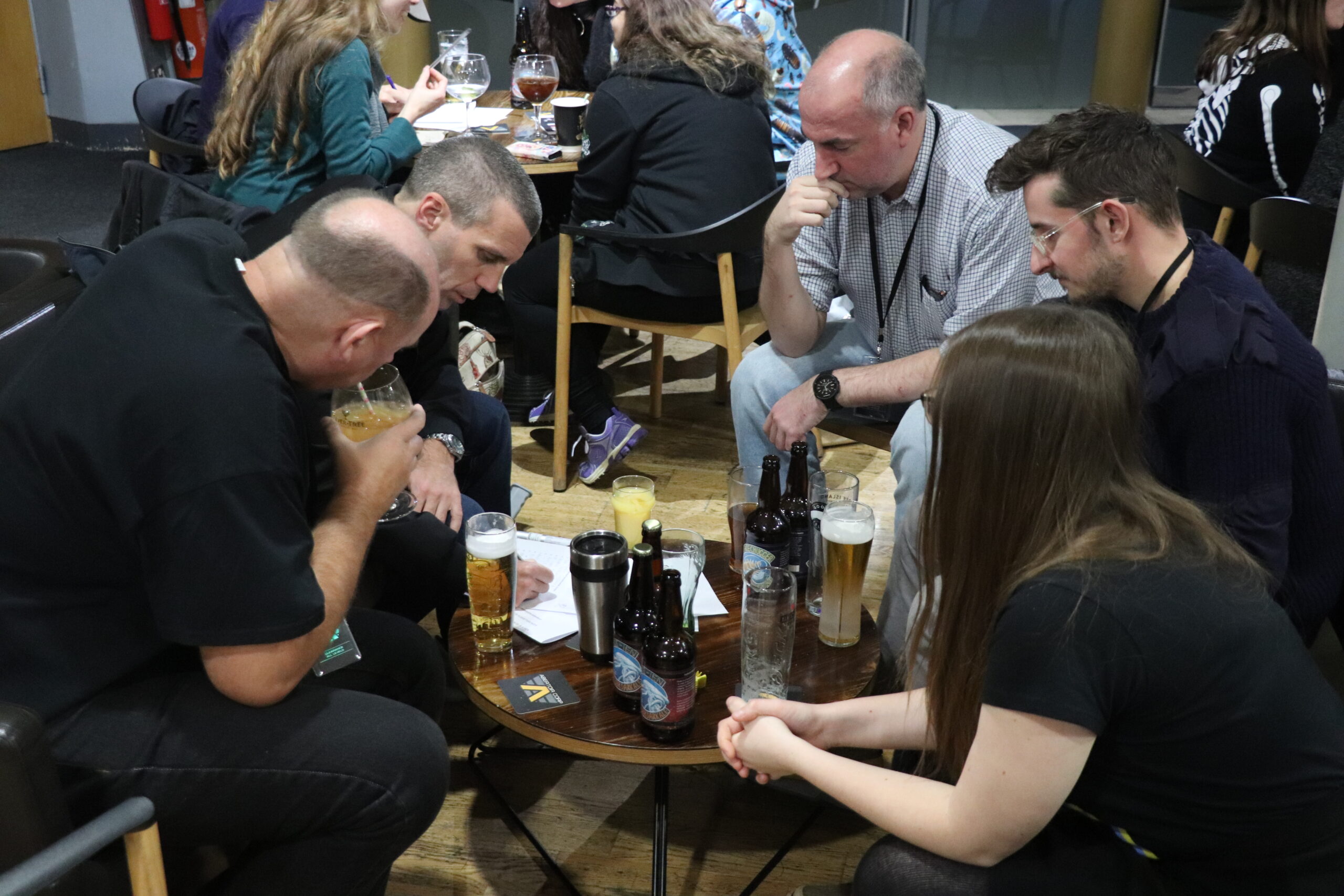 A group of people sitting around a low table, covered in bottles and glasses, ruminating over a sheet of paper.