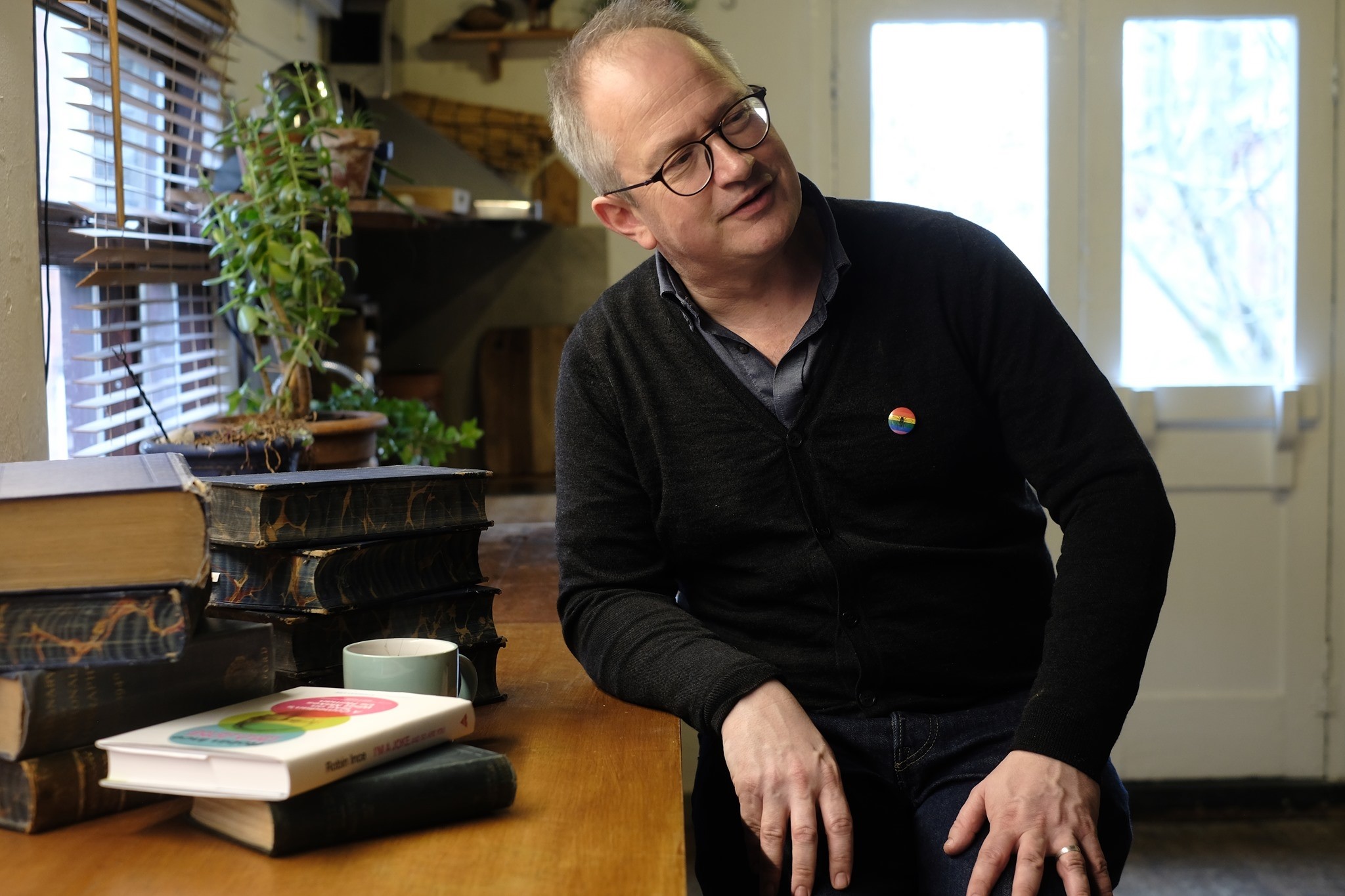 Robin Ince, posing by a desk with books, plants and a cup of tea.