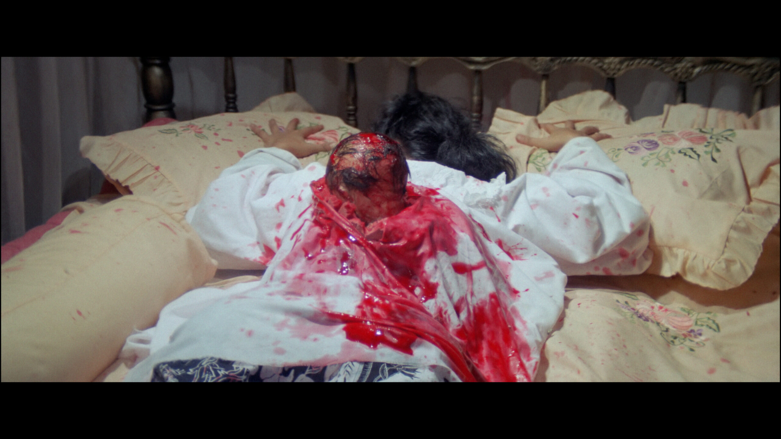 A bloodied body laying on a bed with a head bursting from the back.