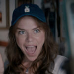 A woman wearing a baseball cap has her mouth open in an excited expression, while holding up a plastic fan.