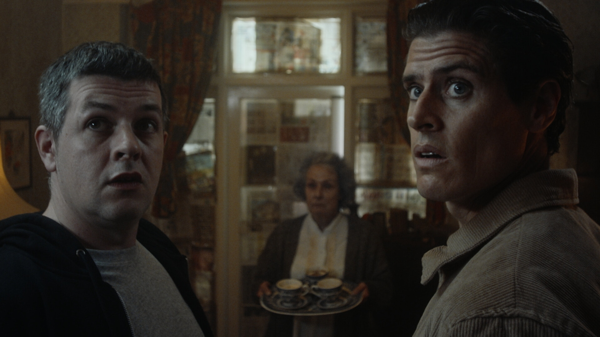 In the foreground, two men face the camera and looking in shock at something out of frame. Between them, in the background, and old woman is holding a tray of tea. The windows behind her are covered in newspaper.