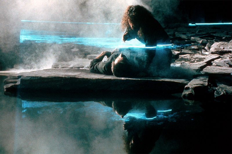 A screen shot from The Keep showing a prone figure, and another leaning over them, on a stone jutting into water. The is mist and a blue light around them.