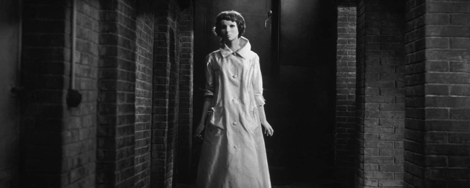 Black and white. A woman wearing a long white coat and a full face mask stands in a long corridor, framed by brick arches.