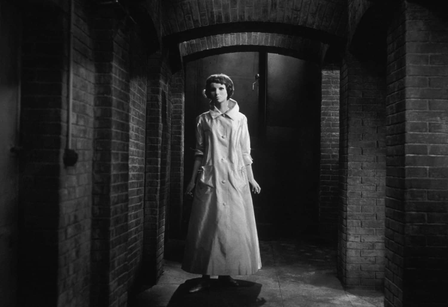 Black and white. A woman wearing a long white coat and a full face mask stands in a long corridor, framed by brick arches.