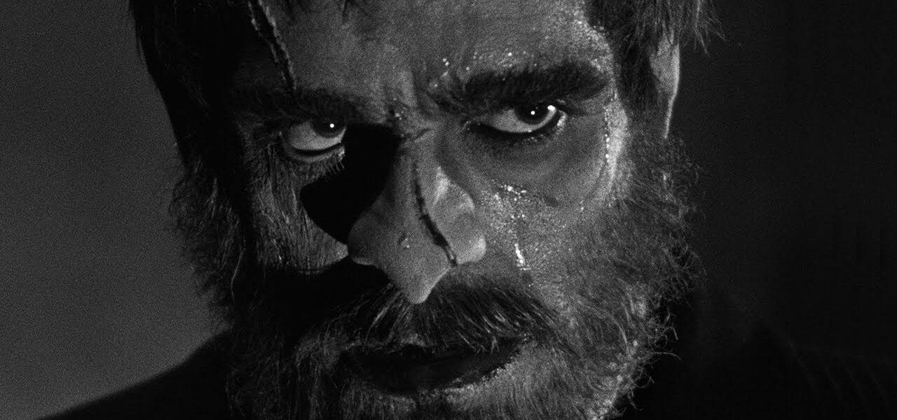 Black and white. An extreme close-up on a rugged man's face. He has scars on his nose and forehead, and a full beard.