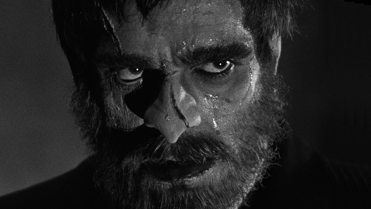Black and white. An extreme close-up on a rugged man's face. He has scars on his nose and forehead, and a full beard.