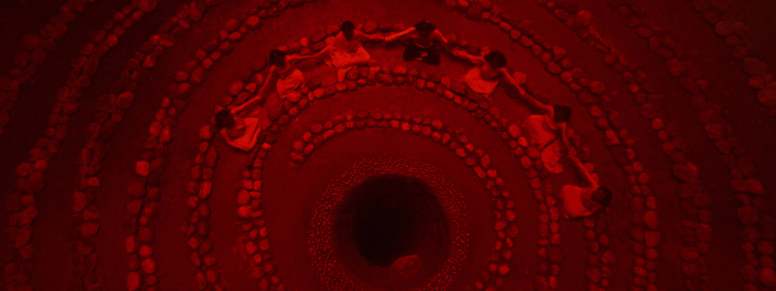 A bird's-eye view of a ritual: stones are arranged in a spiral and seven figures sit, cross-legged, near the centre, where a deep hole is surrounded by smaller stones.