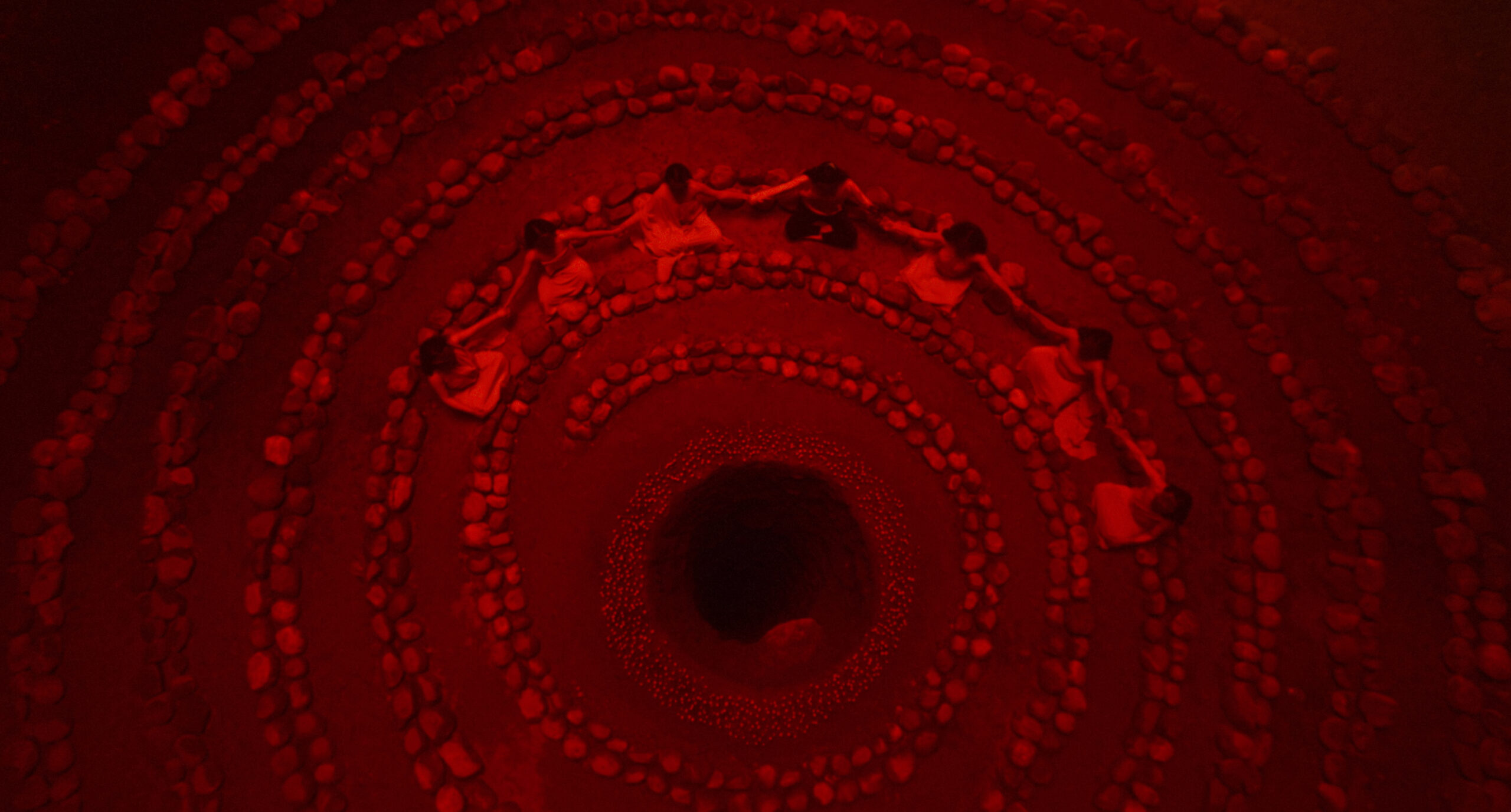 A bird's-eye view of a ritual: stones are arranged in a spiral and seven figures sit, cross-legged, near the centre, where a deep hole is surrounded by smaller stones.