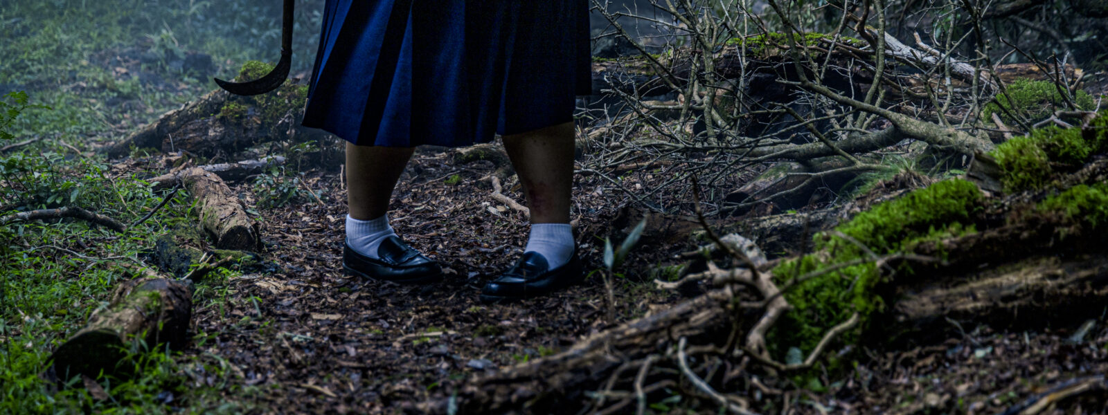 A low-angle view of a forest floor. Framed centrally are the inwardky-pointing feet and legs of a young girl wearing a school uniform, a scythe in her hand at her side. We can only see the lower half of her body.